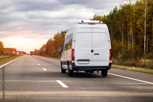 White cargo van driving on highway at sunset, autumn trees line road, delivery vehicle traveling on asphalt route during golden hour with cloudy sky