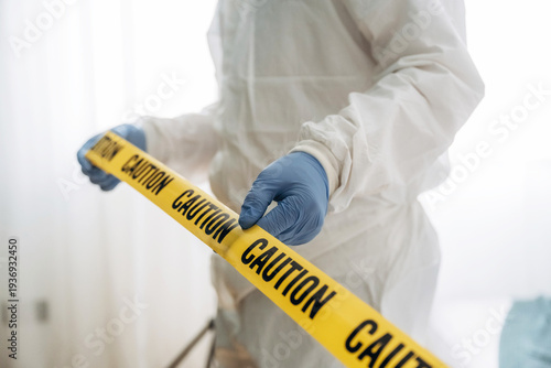 Forensic expert in protective gear holds yellow caution tape in a well-lit indoor setting, indicating a crime scene investigation with blurred background elements