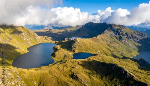 An aerial panorama showcases rugged, mountainous terrain and several placid lakes, framed by clouds in a bright blue sky