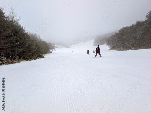 Skiers going down wide groomed ski run lined with trees during foggy weather