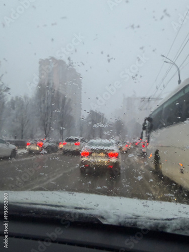 Car driving on road during heavy snow weather. View from inside vehicle through wet windshield with snow and rain drops. Traffic jam in city street during winter storm and cold day.