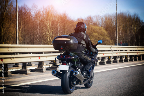 Person rides black sport motorcycle with top case on highway, sun shines, road trip, travel, outdoor adventure, spring weather, blue sky