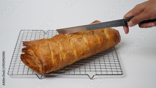 Isolated white hand holding a knife about to cut a whole picnic roll filled with minced beef, boiled eggs, and cheddar cheese wrapped in puff pastry and then baked, placed on a cooling rack.