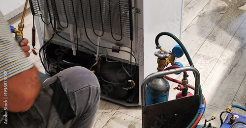Refrigerator repair professional at work, domestic appliance back panel removed, showing compressor, condenser coils, using refrigerant tanks, pressure gauges, hoses, welding torch for service