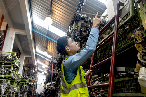 Young Asian male engineer worker with safety vest inspecting at auto spare parts warehouse. Industrial labor man checking and counting product while climbing the ladder surrounded by many engine parts
