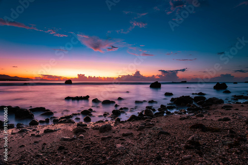 View of a tranquil beach with rocks scattered in the foreground and a vibrant sunset painting the sky with hues of blue, orange, and pink, Mati, Davao Region, Philippines.