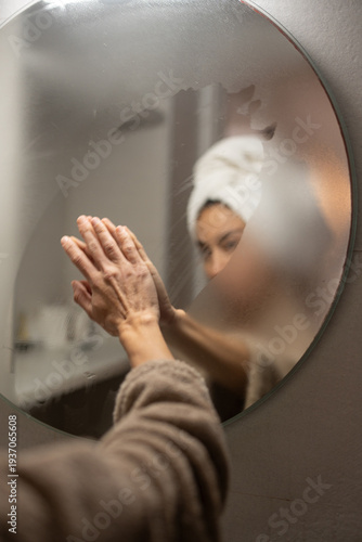 Woman wiping foggy bathroom mirror with hands after shower, soft reflection through steamed glass, towel on head, morning hygiene ritual, self care wellness concept at home