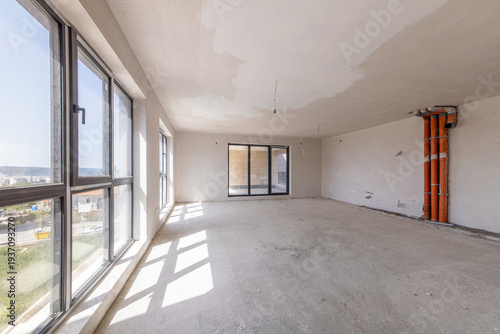 Interior of an empty room under construction in a new apartment. Unfinished space with raw concrete walls, floor, and a large window. Renovation and real estate concept.