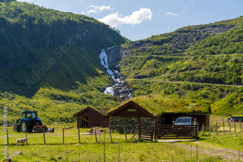 View of emerald hills embrace a cascading waterfall, rustic wooden cabins with grass roofs nestle in the valley, and a blue tractor rests in Holedalen, Vestland, Norway.