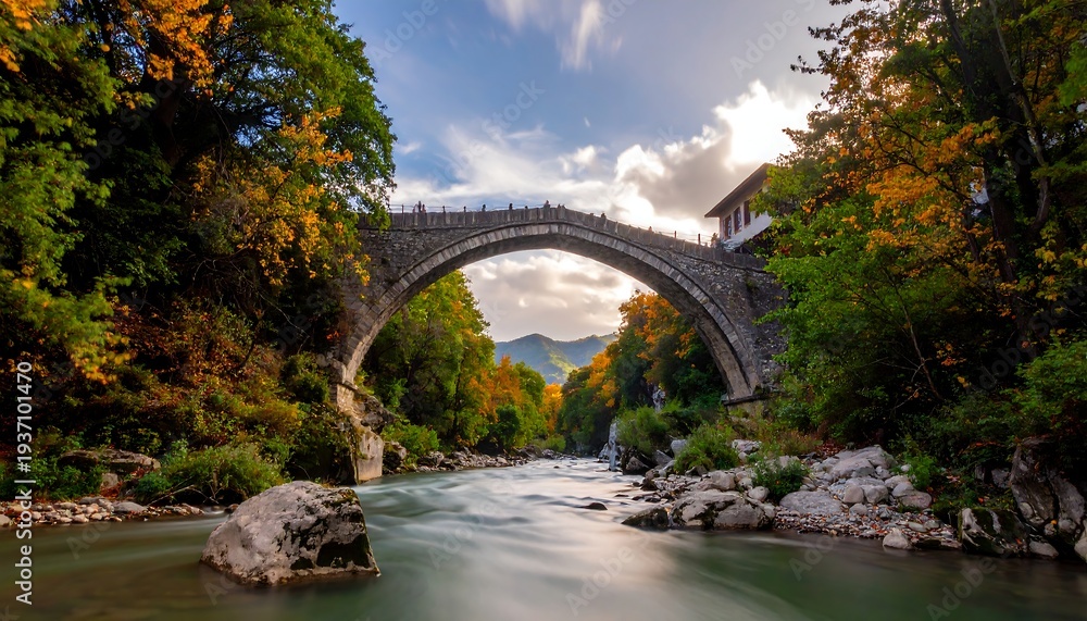 Obraz premium A stone bridge arches over a flowing river, flanked by autumn foliage under a partly cloudy sky