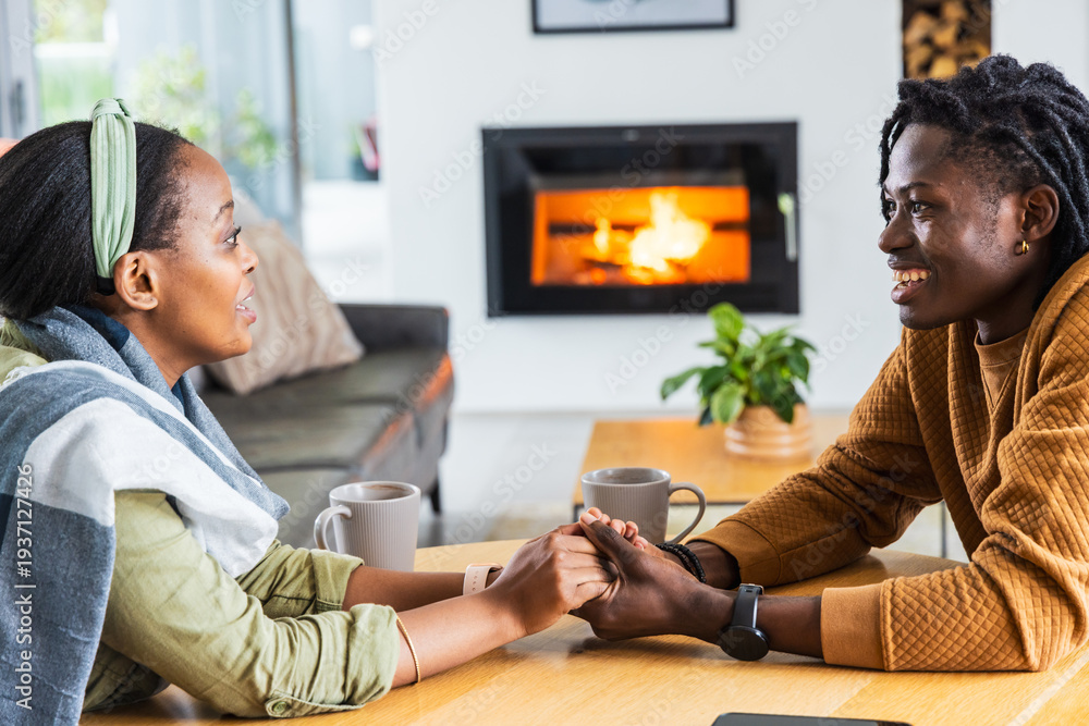 Naklejka premium African American couple holding hands, smiling across wooden table at home with mugs near fireplace