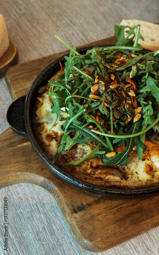 Baked eggs with arugula greens and mixed seeds served in a skillet on a wooden board in a cozy restaurant setting