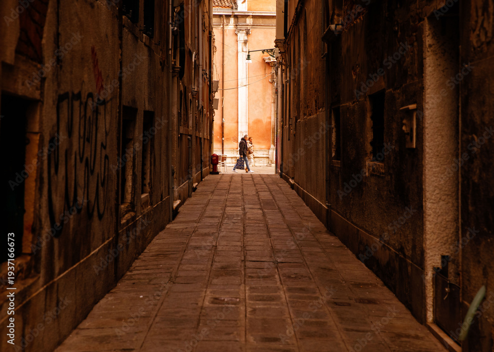 Obraz premium Venice, Italy - February 04, 2024: Narrow alleyway in Venice, Italy, featuring cobblestone pavement and graffiti on old buildings with a couple walking in the distance