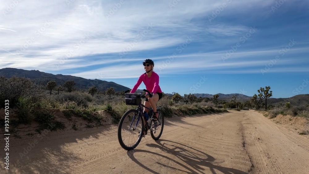 Fototapeta premium Mature woman rides a bike along a dirt path in Joshua Tree National Park during a sunny day with mountains in the background
