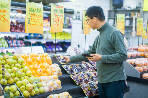 Quadro em tela A middle-aged male customer uses his smartphone while selecting packaged fruits