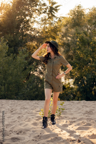 Woman walks on sandy ground in nature during sunset wearing a green outfit and black boots with sunlight shining through trees