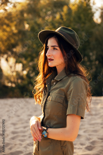 Young woman stands on sandy ground wearing a hat and a green shirt near trees during daytime