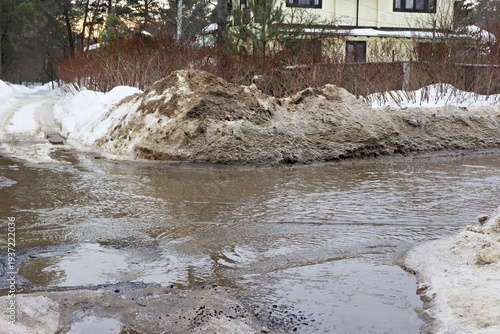 Melting dirty snow piles flank a muddy stream flowing across a road surface near a suburban dwelling