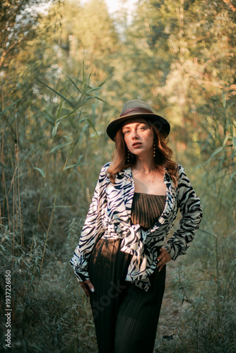 Woman stands in green forest with hat and animal print outfit while touching a branch during daylight in autumn season