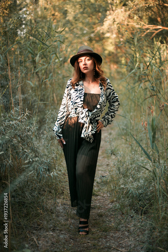 Woman stands in green forest with hat and animal print outfit while touching a branch during daylight in autumn season
