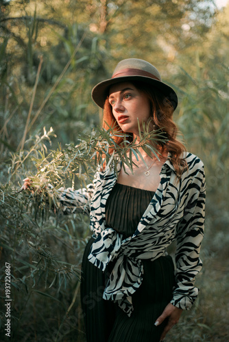 Woman stands in green forest with hat and animal print outfit while touching a branch during daylight in autumn season