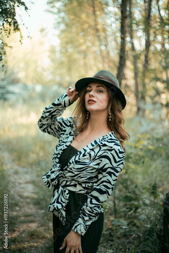Model poses in nature wearing a zebra print top and hat while surrounded by trees on a sunny day