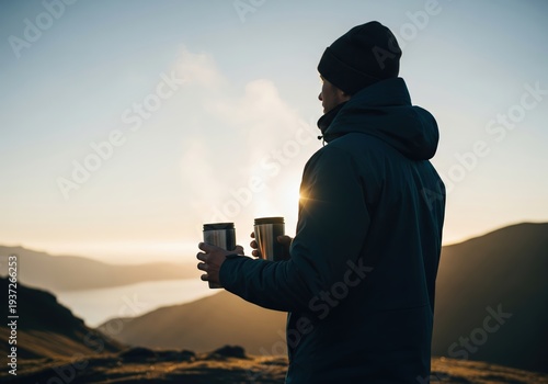 Man stands on mountain holding two cups while watching sunrise over valley and lake in early morning