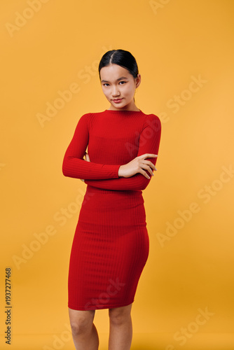 Konstfotografi Young woman in red dress stands with crossed arms looking confidently at camera on yellow background