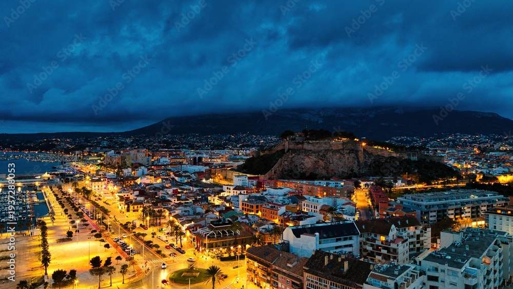 Naklejka premium Aerial drone view of Dénia at twilight with illuminated streets surrounding the historic castle hill. Dramatic clouds cover Montgó mountain while the Mediterranean harbor and city lights create a cine
