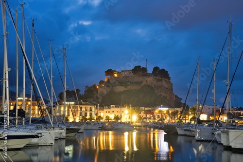 Marina harbor in Dénia on the Mediterranean coast of Spain filled with sailboats and yachts. The historic castle hill rises above the waterfront beneath dramatic sunset clouds reflected in calm water.