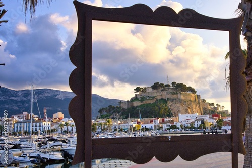 Framed view of the marina harbor and historic castle hill in Dénia, Spain at golden sunset. Sailboats and waterfront promenade create a cinematic Mediterranean coastal cityscape under dramatic clouds.