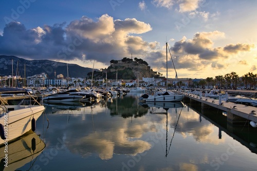 Harbor filled with sailboats in Dénia on the Mediterranean coast of Spain with the historic castle hill rising above the town. Golden sunset clouds create dramatic light reflected in calm harbor water