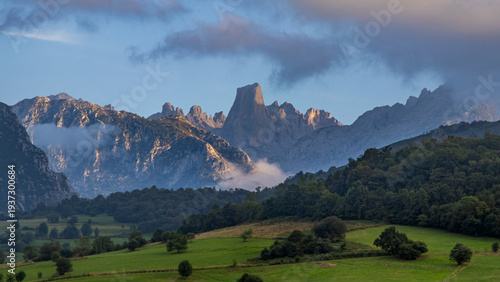 Tableau sur toile Majestuosa vista del Naranjo de Bulnes en los Picos de Europa, con sus paredes calizas iluminadas por el sol poniente sobre un valle verde y bosques envueltos en jirones de niebla