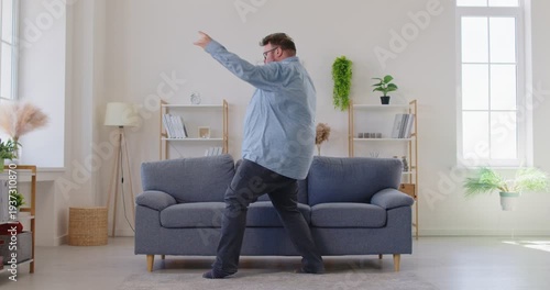 Expressive fat overweight man in glasses dancing and singing with upbeat energy near a sofa in living room, enjoying music and the moment at home, carefree. Obese male person having fun indoors.