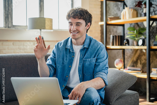 Happy young man makes videocall seated on sofa at home, greets friend or family start video conference using application on laptop. Virtual meeting event, tech, remote talk concept