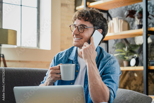 Remote work, laptop, phone and man on sofa with drink, conversation at home. Male person, small business and technology in living room for contact, ecommerce and interior design on internet