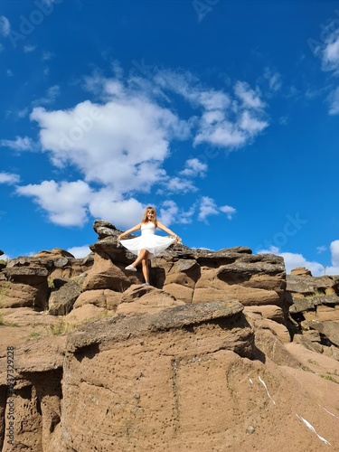 Young woman in white dress stands on rocky terrain under a blue sky with clouds, showcasing natural landscape and outdoor beauty in a scenic location