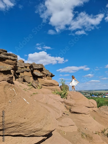 Young girl in white dress stands on rocky outcrop against blue sky with clouds, overlooking green valley and distant village in the background