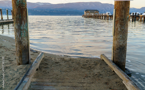 A small sandy beach access path leads between two weathered wooden dock posts toward the calm waters of a lake. In the distance, a pier and boathouse extend into the lake with mountains rising