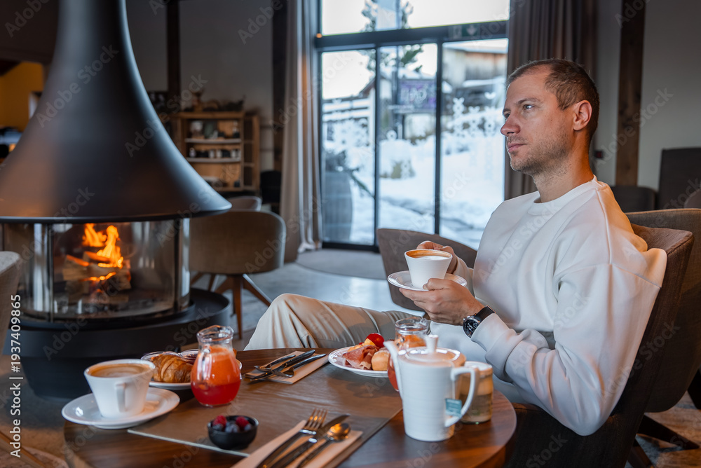 Naklejka premium A guest in a light sweater sips coffee by a modern fireplace in a Dolomites boutique hotel, with croissants, berries, cured meats, and snow beyond large glass doors.