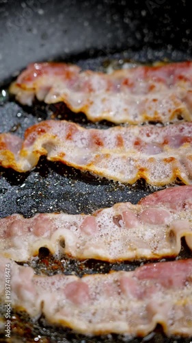 Tongs flip sizzling bacon strips in pan, home chef adjusts placement, edges crisping and browning, oil bubbling and spattering, closeup cooking action