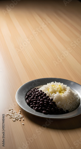 Simple serving of rice and black beans on a plain metal plate placed in the lower right corner of a large light wooden table, most of the table left empty as negative space.