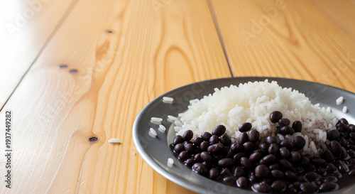 Simple serving of rice and black beans on a plain metal plate placed in the lower right corner of a large light wooden table, most of the table left empty as negative space.