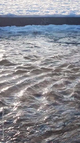 Vertical close-up shot of flowing clean shallow river or stream water surface with sunbeams and frozen snowy sand shore at sunny winter or early spring day. Aquatic texture pattern natural background.