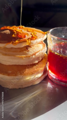 Close-up shot of honey being drizzled over stacked pancakes, with a glass of honey syrup in the background, creating a delicious breakfast scene.
