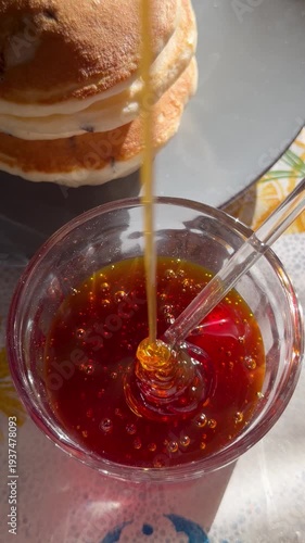 Close-up of honey being poured into a glass jar with pancakes in the background. Perfect for food content, breakfast cooking, and food styling videos.
