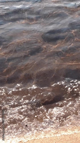 Vertical close-up shot of clean flowing waves of shallow river or stream water surface and frozen ice-covered sand shore at sunny winter or pring day. Aquatic texture pattern natural background.