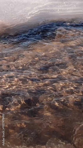 Vertical close-up shot of flowing clean shallow river or stream water surface and frozen ice-covered shore at sunny winter or early spring day. Aquatic and icy texture pattern natural background.