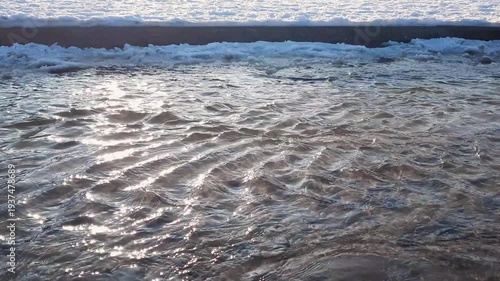 Close-up of flowing clean shallow river or stream water surface with sunbeams and frozen snow-covered sand shore at sunny winter or early spring day. Aquatic texture pattern natural background.