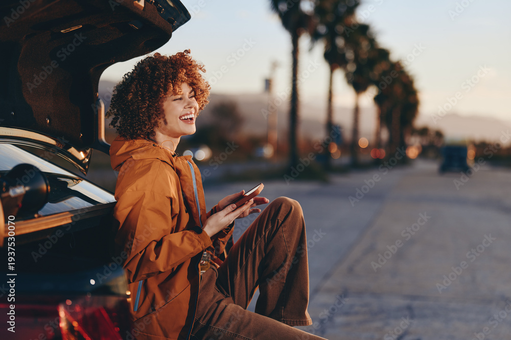 Fototapeta premium Young woman with curly hair wearing rainbow sweater sits on car trunk smiling and holding smartphone outdoors on sunny day with palm trees and road in background.